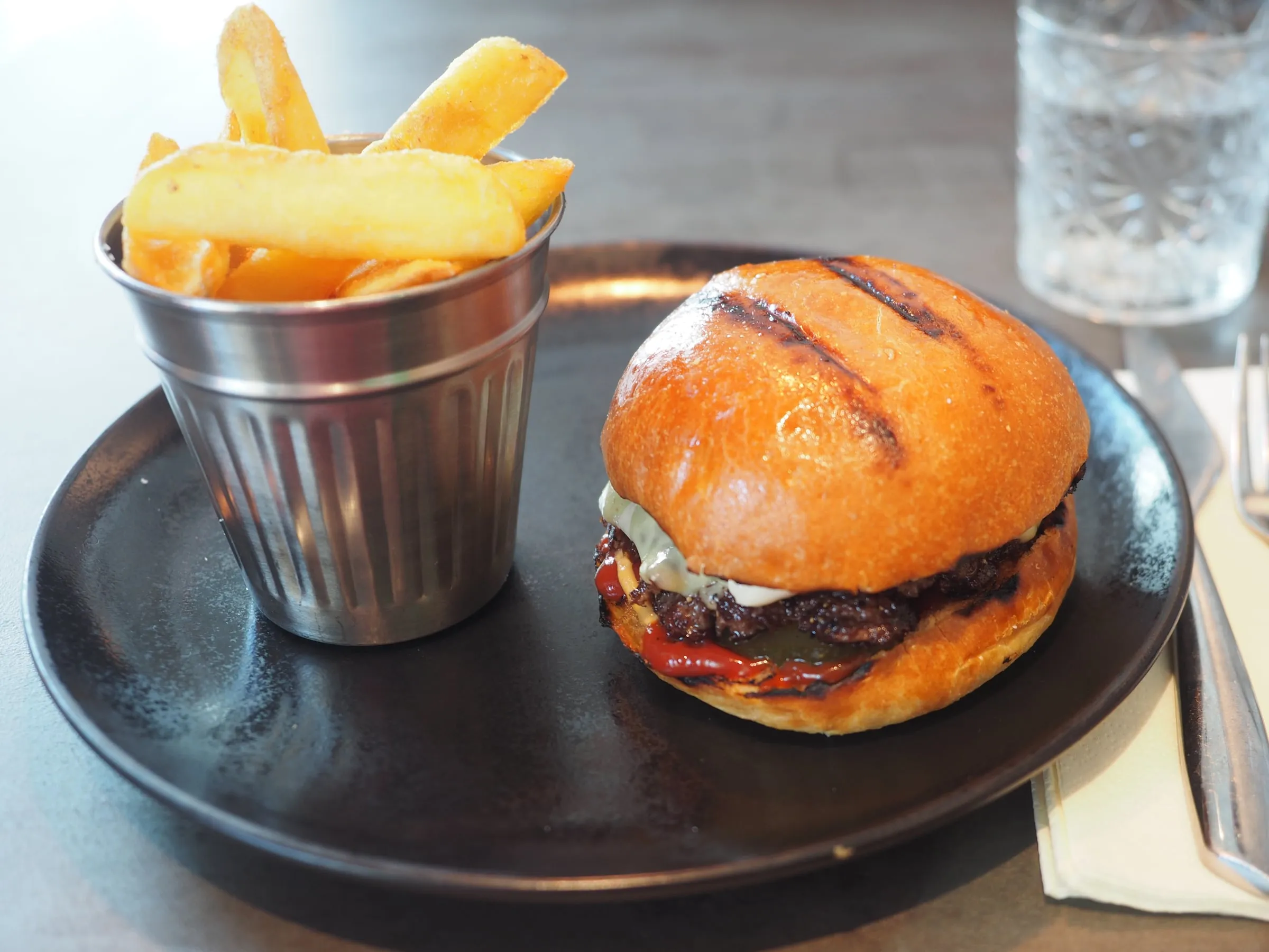 Smash burger con carne de orillas doradas, queso, pepinillo y cátsup en pan brioche marcado a la parrilla, con papas fritas