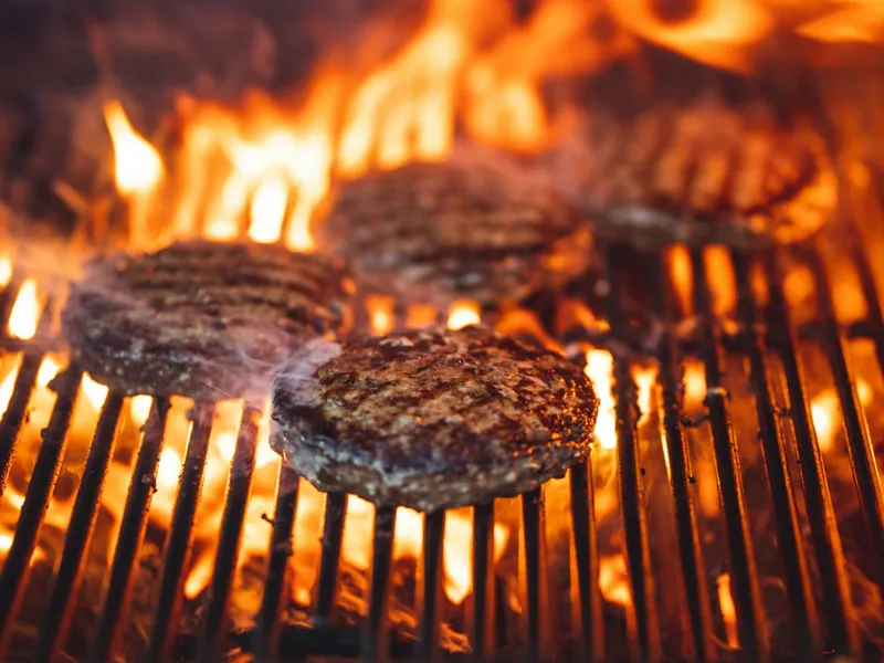 Burger patties searing over a hot charcoal fire, building a dark crust before reaching a safe internal temp of 160°F (71°C)