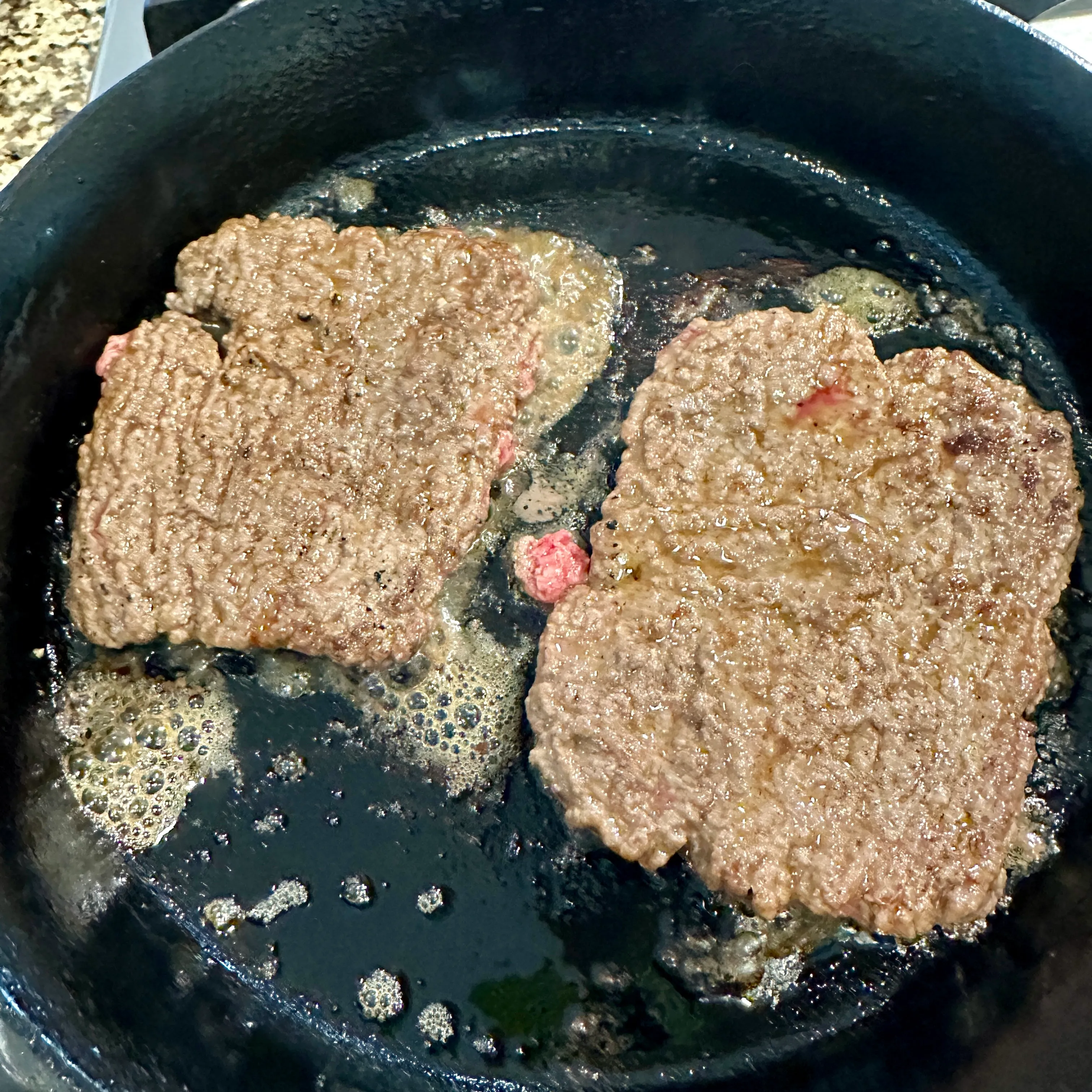 Thin pressed ground beef patties searing in a hot cast-iron skillet for a homemade patty melt