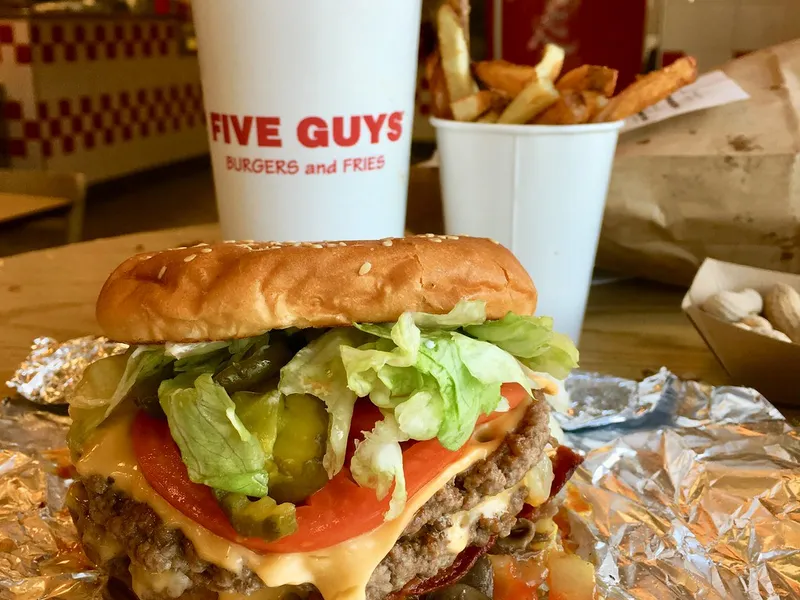 Five Guys Bacon Cheeseburger at the restaurant: two patties with melted American cheese, crispy bacon, tomato, pickles and lettuce on a toasted sesame bun, open on its foil with fries