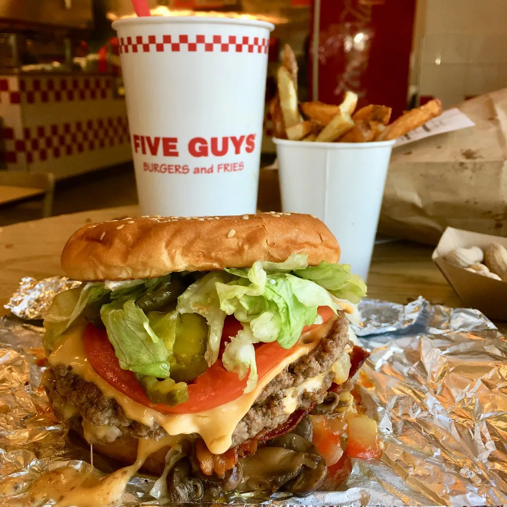 Five Guys Bacon Cheeseburger at the restaurant: two patties with melted American cheese, crispy bacon, tomato, pickles and lettuce on a toasted sesame bun, open on its foil with fries