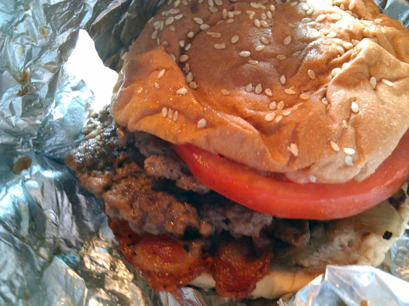 Five Guys bacon burger with no cheese half unwrapped from its foil: crisp bacon strips on well-done beef patties, tomato and a toasted sesame bun
