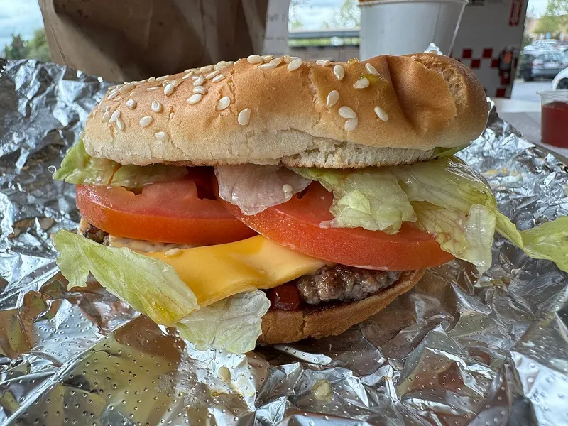 Hamburguesa con queso pequeña de Five Guys con una sola carne, queso americano, tomate y lechuga en pan de sésamo, abierta sobre su papel de aluminio, modelo de esta receta casera