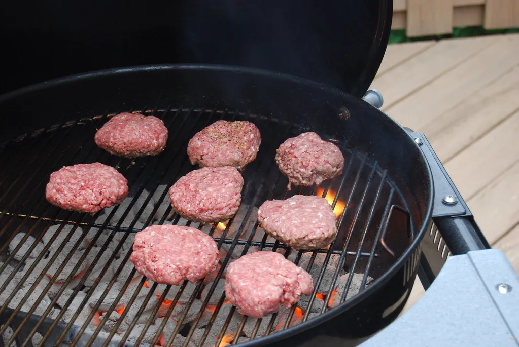 Seasoned raw beef patties on a hot charcoal kettle grill for a charbroiled cheeseburger