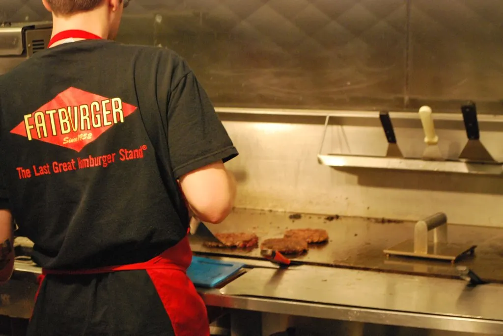 Cuisinier qui aplatit des steaks hachés sur une plaque en inox, une presse à burger posée à côté