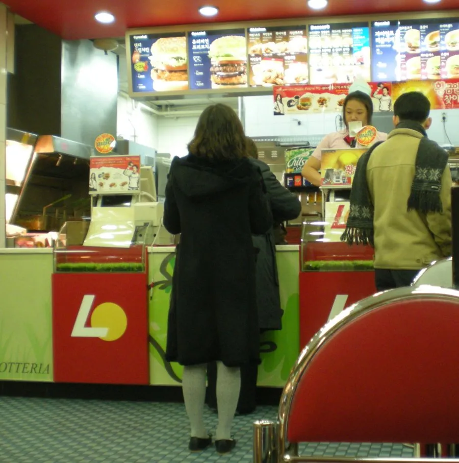 Customers ordering at a Lotteria counter in South Korea under backlit menu boards