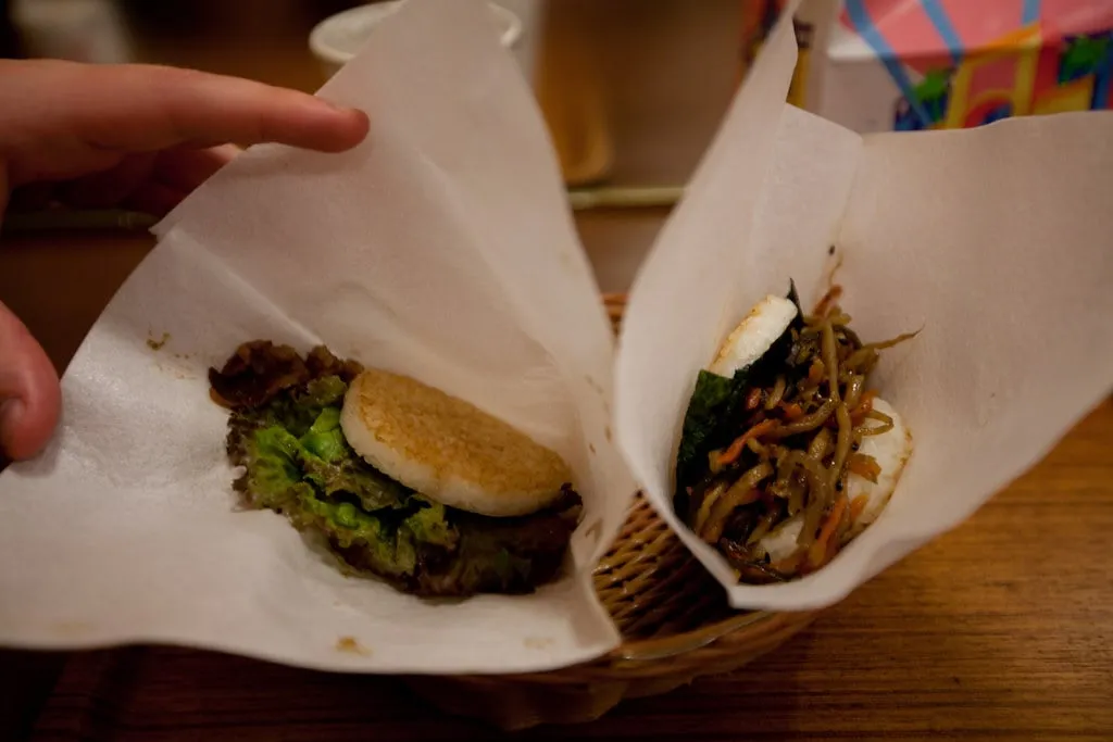 Deux rice burgers MOS dans leur papier, l’un garni de bœuf grillé et de salade, l’autre de kinpira de bardane et carotte, entre deux galettes de riz pressé