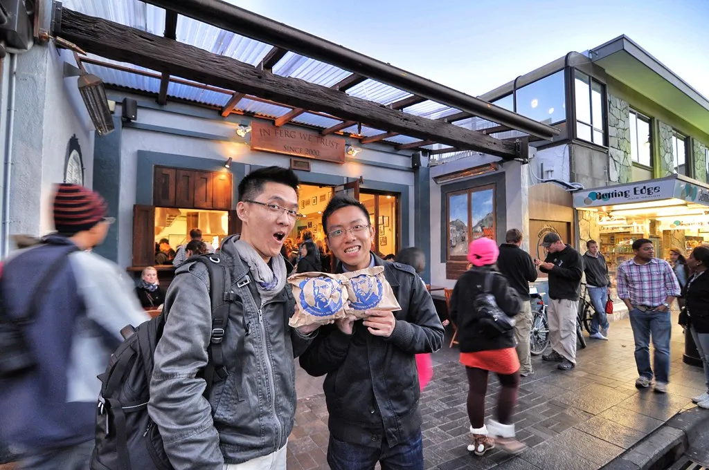 Two visitors holding Fergburger paper bags with the bearded Ferg logo outside the Shotover Street terrace