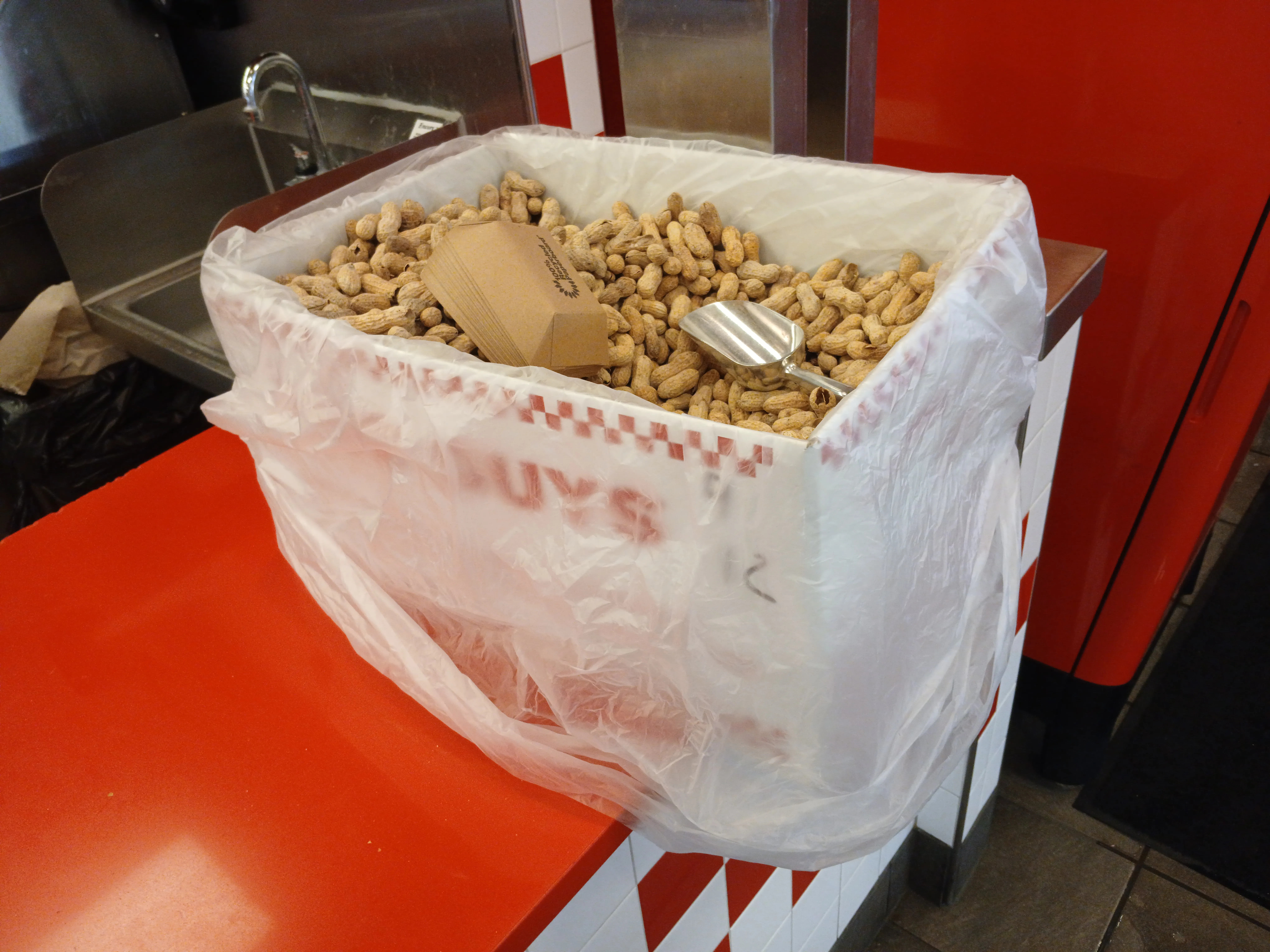 Box of free in-shell peanuts with a scoop and paper trays on a red Five Guys counter, a peanut allergy risk at Five Guys